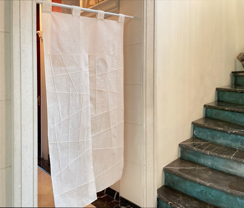 Op de afbeelding: Een wit linnen gordijn met een geometrisch patroon hangt in een deuropening. Het gordijn bestaat uit twee panelen, elk met een ander patroon. Het gordijn hangt aan een witte stang.
