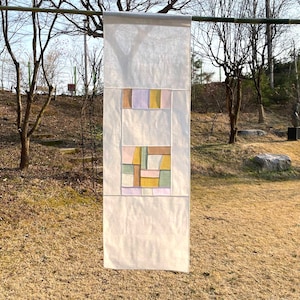 May include: A white fabric wall hanging with a geometric design in shades of pink, yellow, green, and brown.