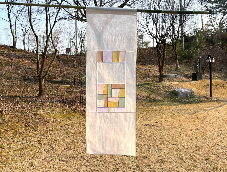 May include: A white fabric wall hanging with a geometric design in shades of pink, yellow, green, and brown.
