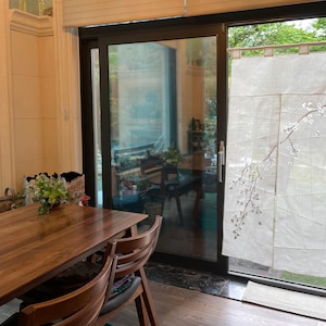 Op de afbeelding: Een houten eettafel met vier stoelen, geplaatst voor een schuifdeur van glas met een wit transparant gordijn met een delicaat bloemenmotief. Het gordijn is gedeeltelijk opengetrokken, waardoor een uitzicht op een weelderige groene tuin zichtbaar is.