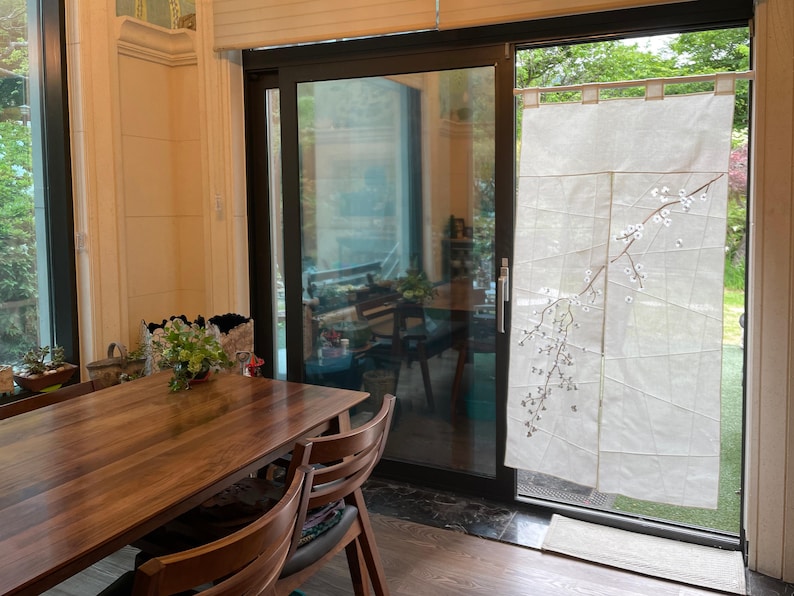 Op de afbeelding: Een houten eettafel met vier stoelen, geplaatst voor een schuifdeur van glas met een wit transparant gordijn met een delicaat bloemenmotief. Het gordijn is gedeeltelijk opengetrokken, waardoor een uitzicht op een weelderige groene tuin zichtbaar is.