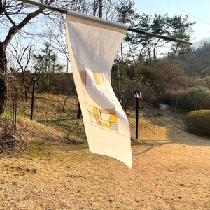 May include: A white fabric banner with a geometric design in shades of yellow, orange, and green. The banner is hanging from a wooden pole in a garden setting.