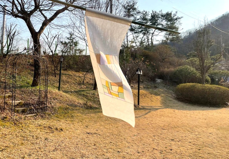 May include: A white fabric banner with a geometric design in shades of yellow, orange, and green. The banner is hanging from a wooden pole in a garden setting.
