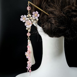 May include: A decorative hair accessory featuring pink flower accents, pearls, and a white tassel. The hair piece has a gold-colored pin and dangling pink beads. The flowers are a soft pink color, and the pearls are white.