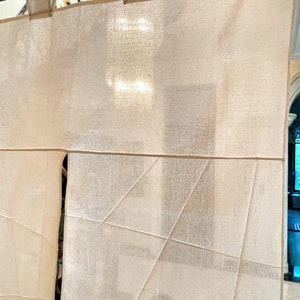 Op de afbeelding: Een wit katoenen gordijn met een geometrisch stikselpatroon. Het gordijn hangt aan een metalen stang.