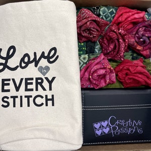 May include: A white canvas tote bag with the text "Love in Every Stitch" printed in black. The bag is filled with fabric scraps in shades of red and green. A black box with the text "Creative Passions" in purple is also included.