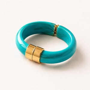 Puede incluir: Un brazalete turquesa con detalles metálicos dorados. El brazalete tiene una forma sólida y redondeada con un acabado brillante. Los detalles dorados están en el cierre y en la parte delantera. Joyería.