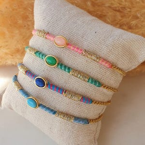 Peut inclure: Cinq bracelets colorés avec des accents dorés et des pierres précieuses de forme ovale. Les bracelets sont empilés sur une surface en tissu beige.