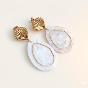 May include: A pair of gold-colored earrings with a seashell design at the top. The earrings have a white oval shape with a gold-colored outline and a marbled effect. The earrings are designed for pierced ears.