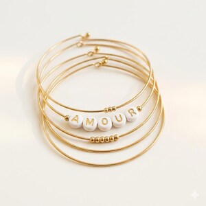 Op de afbeelding: Goudkleurige metalen armbanden met witte letterkralen met de tekst "AMOUR". De armbanden zijn gestapeld, met de woordkralen gecentreerd op een van de armbanden. Kleine gouden kralen accentueren het ontwerp.