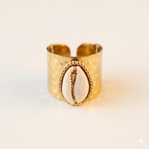 May include: A gold-coloured adjustable ring with a hammered texture. The ring features a white and tan seashell in an oval setting, surrounded by small gold beads. The ring is set against a white background.