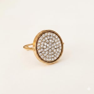 Pode incluir: Anel dourado com uma grande face circular densamente coberta por pequenos cristais transparentes. O anel tem uma banda simples e uma moldura elevada ao redor da face coberta de cristal, criando um design elegante e brilhante.