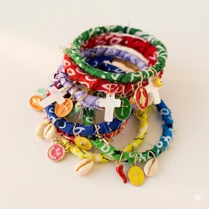 Könnte beinhalten: Eine Ansammlung von stoffummantelten Armbändern in verschiedenen Farben, darunter Grün, Rot, Blau und Gelb. Jedes Armband ist mit Anhängern wie Kreuzen, Muscheln und religiösen Symbolen verziert. Die Armbänder liegen auf einem weißen Hintergrund.