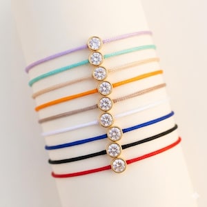 Può includere: Una collezione di braccialetti di filo colorati con una lunetta dorata che incastona una gemma rotonda e trasparente. I braccialetti sono disponibili in vari colori, tra cui rosso, nero, blu, bianco, marrone, arancione, turchese e viola.