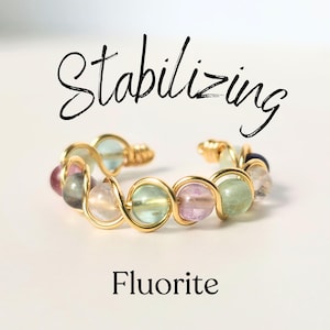 May include: A gold wire wrapped ring with a variety of fluorite beads in shades of green, pink, and purple. The ring is labeled "Stabilizing Fluorite".