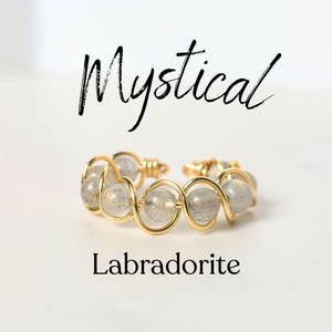 May include: A gold-colored ring with clear, gray, and iridescent labradorite beads. The ring is open and adjustable. The word "Mystical" is written above the ring, and the word "Labradorite" is written below it.