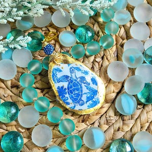 May include: A gold and blue sea turtle charm pendant with a white and blue floral design. The pendant is hanging from a gold chain and is surrounded by blue and white glass beads.
