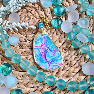 May include: A blue and pink patterned key chain with a gold-colored metal ring and a blue and white beaded charm. The key chain is shaped like a seashell and is surrounded by blue and white glass beads.