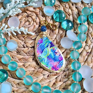 May include: A gold-leafed, resin-coated keychain featuring a colorful sea turtle design. The keychain is surrounded by blue and white glass beads on a woven straw surface.