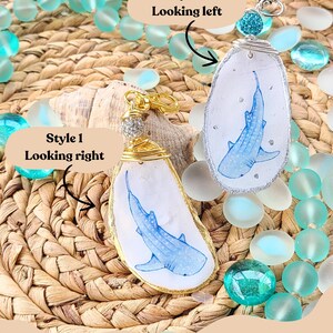 May include: Two handmade shark-themed keychains. One has a gold border, the other a silver border. Each features a blue shark illustration on a white background. The keychains are displayed with turquoise beads and a woven background.