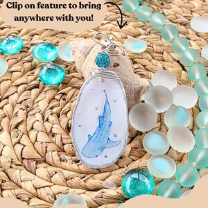 May include: Handmade oyster shell keychain featuring a blue whale shark design. The shell is outlined with silver glitter and has a clip-on feature. The background includes turquoise glass beads and a woven texture. Text reads: "Clip on feature to bring anywhere with you!"