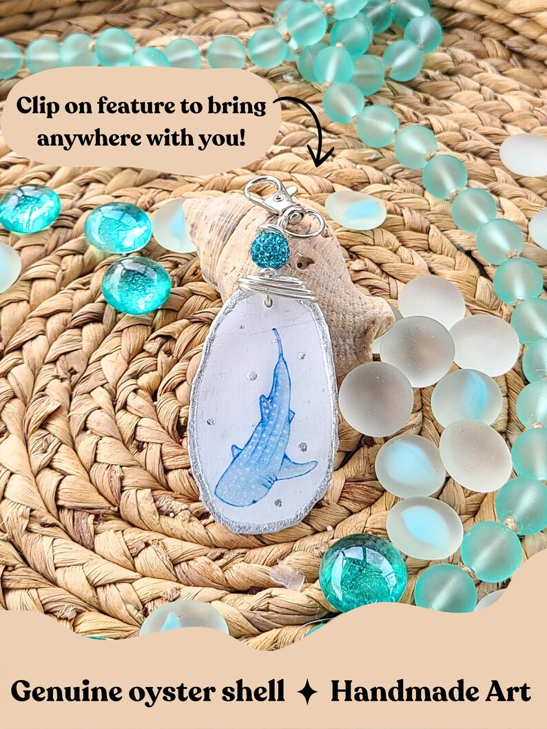 May include: Handmade oyster shell keychain featuring a blue whale shark design. The shell is outlined with silver glitter and has a clip-on feature. The background includes turquoise glass beads and a woven texture. Text reads: "Clip on feature to bring anywhere with you!"
