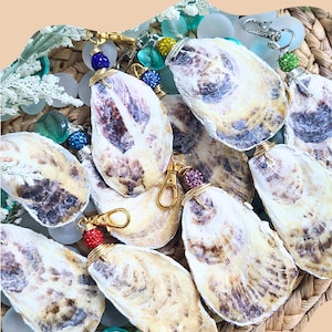 May include: A collection of oyster shell keychains with gold-colored hardware and colorful beaded accents. The shells vary in color and texture, with text that reads: "Examples of what the backs of each oyster piece looks like. No oyster shell is the exact same."