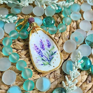 May include: A gold-painted, oval-shaped pendant with a hand-painted lavender flower design. The pendant is attached to a gold chain with a lobster clasp. The pendant is surrounded by light blue glass beads.
