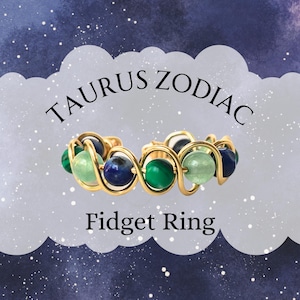 May include: A gold-colored fidget ring with green, blue, and clear beads. The ring is labeled "TAURUS ZODIAC Fidget Ring" against a starry, blue and purple background.