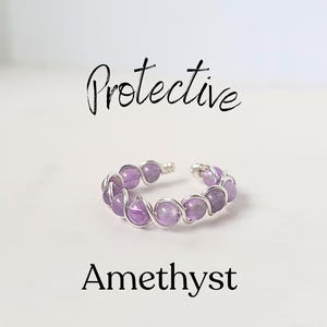 May include: A silver wire wrapped ring with amethyst stones. The ring is on a white background and the text "Protective Amethyst" is written below the ring.