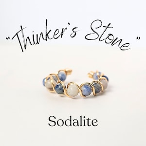 May include: A gold wire wrapped ring with blue and white sodalite beads. The ring is on a white background. The text "Thinker's Stone" is above the ring and the text "Sodalite" is below the ring.