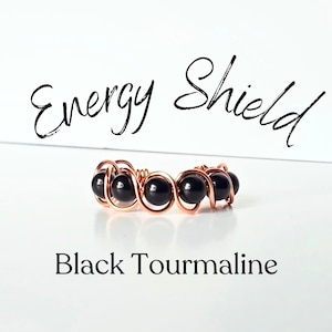 May include: A ring featuring a series of round, polished black tourmaline beads secured within a continuous, spiraling copper wire frame. The text "Energy Shield" appears above the ring, and "Black Tourmaline" is printed below the ring.