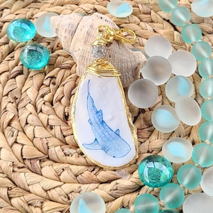 May include: A handmade pendant featuring a blue whale shark illustration on a white background, framed in gold wire. The pendant is attached to a gold-colored clasp and adorned with a crystal bead. The pendant rests on a seashell, surrounded by teal and clear glass beads.