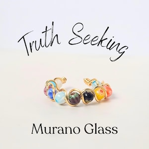 May include: A gold wire ring with colorful Murano glass beads. The ring is made with a twisted wire design and features a variety of colors, including blue, green, yellow, and red. The text "Truth Seeking" is written above the ring and "Murano Glass" is written below the ring.