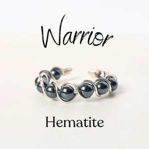 May include: A silver wire ring with black hematite beads. The ring is on a white background and the word "Warrior" is written above the ring. The word "Hematite" is written below the ring.