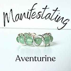 May include: A wire-wrapped ring featuring round, light green aventurine beads secured within a silver-toned metal coil. The text "Manifestating" appears above the ring, and the word "Aventurine" is printed below the jewelry piece.