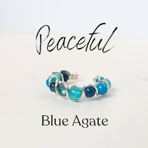 May include: A silver wire wrapped ring with blue agate stones. The ring is on a white background and the word "Peaceful" is written above the ring. The words "Blue Agate" are written below the ring.