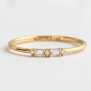 May include: A slender gold ring with a fine band. The ring is adorned with three baguette-cut diamonds at the centre, with two smaller round diamonds on either side. The ring is set against a white backdrop.