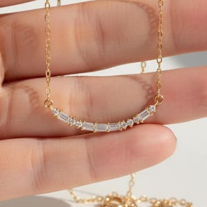 Könnte beinhalten: Eine zarte goldene Halskette mit einem gebogenen Anhänger. Der Anhänger ist mit einer Reihe kleiner, klarer, rechteckiger und runder Edelsteine verziert. Die Halskette wird in einer Hand gehalten und zeigt ihre feine Kette und ihren Verschluss.