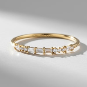 Könnte beinhalten: Ein zarter Goldring mit kleinen Diamanten und Baguette-Schliff-Steinen. Der Ring liegt auf einer weißen Oberfläche, mit weichen Schatten. Ein feines Schmuckstück.