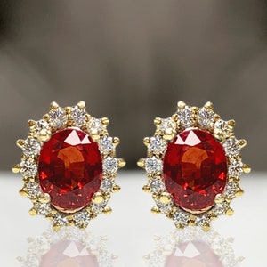 May include: A pair of gold earrings with a red oval gemstone surrounded by a halo of smaller white gemstones.