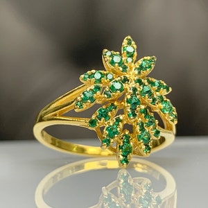 May include: A gold ring with a green gemstone design. The ring features a stylized leaf pattern with multiple green gemstones set in the gold.