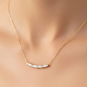May include: A gold necklace with a delicate chain, adorned with a row of rectangular, clear gemstones. The gemstones are set in a curved line, creating a subtle sparkle. The necklace is a simple yet elegant piece of jewellery, perfect for everyday wear.
