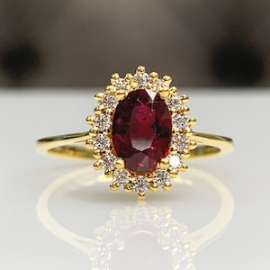 May include: A gold ring featuring a large oval-cut ruby gemstone surrounded by a halo of small diamonds. The ring's band is a simple, polished gold. The ruby is a deep red color, and the diamonds sparkle.