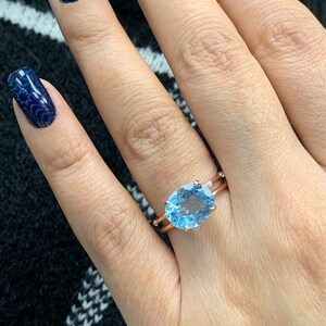 May include: A rose gold ring with a large oval-shaped blue topaz gemstone. The ring has a delicate band with small diamonds on either side of the gemstone.