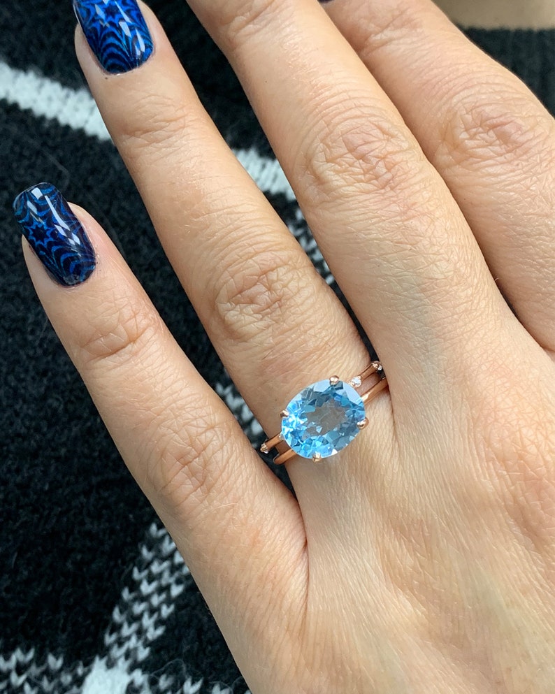 May include: A rose gold ring with a large oval-shaped blue topaz gemstone. The ring has a delicate band with small diamonds on either side of the gemstone.