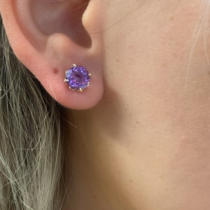 Pu&ograve; includere: Un paio di orecchini a perno d'oro con una pietra preziosa viola al centro.