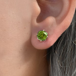 Pu&ograve; includere: Un paio di orecchini con pietre preziose verdi con una montatura in argento. Gli orecchini sono piccoli e rotondi e sono incastonati in una montatura a griffe.