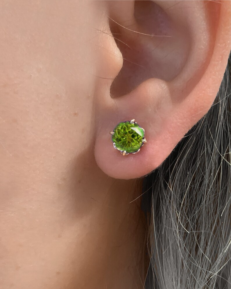 Pu&ograve; includere: Un paio di orecchini con pietre preziose verdi con una montatura in argento. Gli orecchini sono piccoli e rotondi e sono incastonati in una montatura a griffe.
