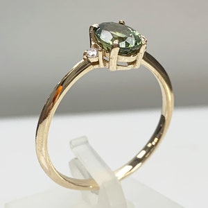 May include: A gold ring featuring a green oval gemstone and a smaller diamond accent. The ring is displayed on a white stand.